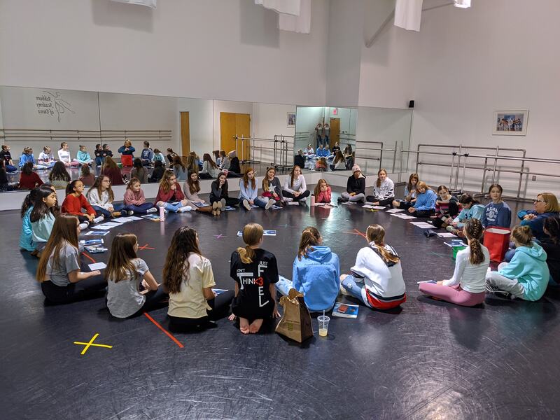 Twenty six young dance students and their dance teacher sitting in a circle in a dance classroom participating in a leadership lesson.