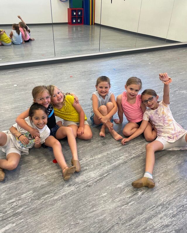 Six young female dance students sitting on the floor, hugging each other, and smiling at the camera.