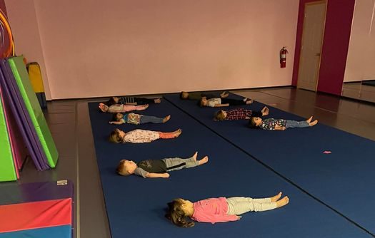Ten toddler-aged kids lying on their back in a row on a blue gymnastics mat looking up at the ceiling.